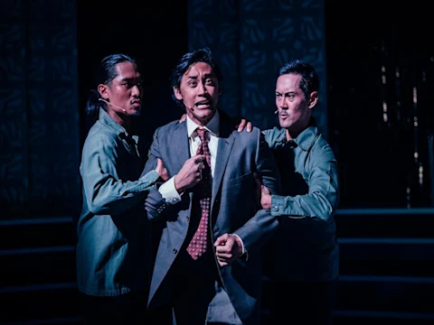 Three men on stage: two in blue shirts hold a man in a suit and tie, who looks distressed, under dramatic lighting with a dark background.