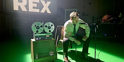 A man in a shirt and tie sits at a microphone beside a reel-to-reel tape recorder in a studio with "REX" projected on the wall behind him.