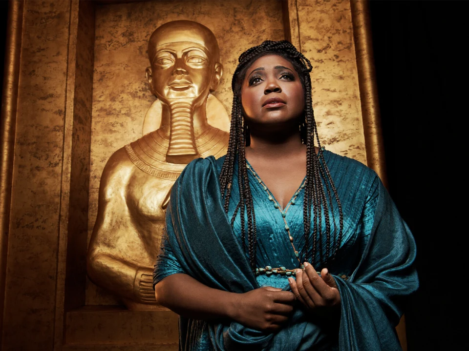 Product media of Aida in New York, showing a woman in teal clothing stands in front of a golden statue. She has braided hair and gazes upward with a solemn expression.