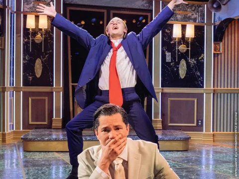 A man in a blue suit and red tie performs energetically on stage while another man in a beige suit covers his mouth in surprise or amusement in the foreground.