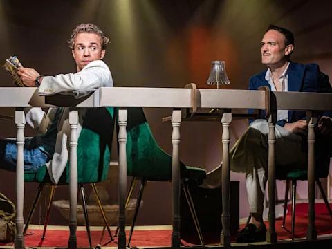 Two men sit at a table with green chairs under dramatic lighting; one looks back holding something, while the other sits facing forward, watching him.