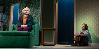 An older woman sits on a green armchair while a younger woman sits on the floor against a wall in a room with patterned wallpaper and bright lighting.