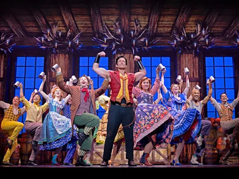 A group of performers in colorful costumes dances and raises mugs on a wooden stage, with a muscular man posing at the center.