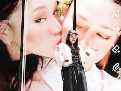 A woman in a white fur jacket and black dress poses with arms raised in front of a large screen showing a close-up image of two faces.