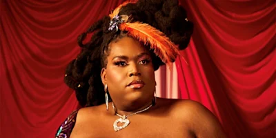 A person with long styled hair, wearing a feathered headpiece, bold makeup, and a heart-shaped necklace poses in front of red draped curtains.