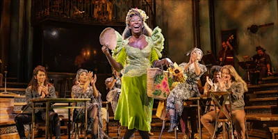A woman in a bright green dress stands smiling with baskets on stage while several people seated at tables applaud. Musicians perform in the background.
