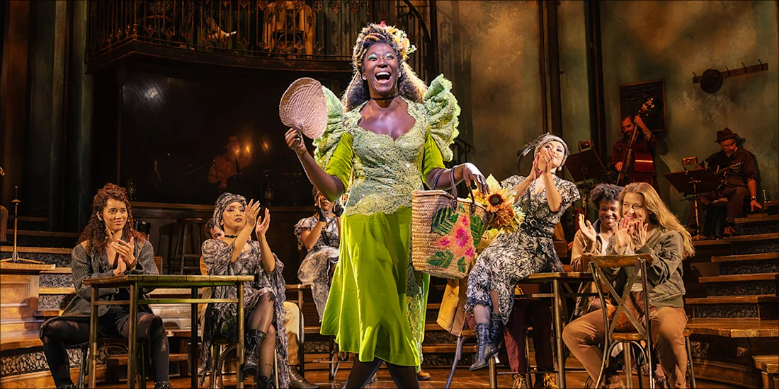 A woman in a bright green dress stands smiling with baskets on stage while several people seated at tables applaud. Musicians perform in the background.