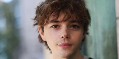 A young person with wavy brown hair and fair skin looks directly at the camera, standing near a green wall with a blurred outdoor background.