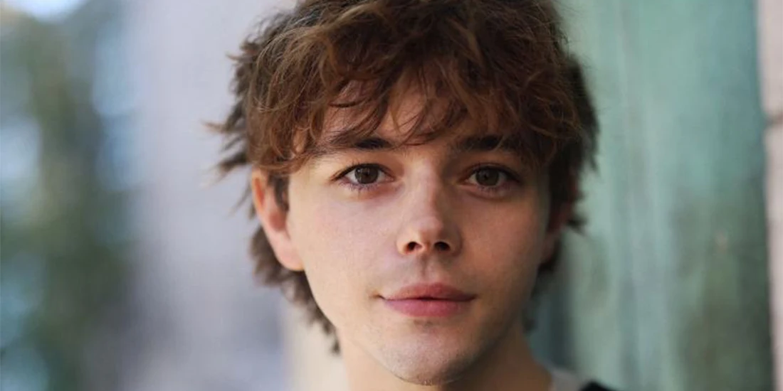 A young person with wavy brown hair and fair skin looks directly at the camera, standing near a green wall with a blurred outdoor background.