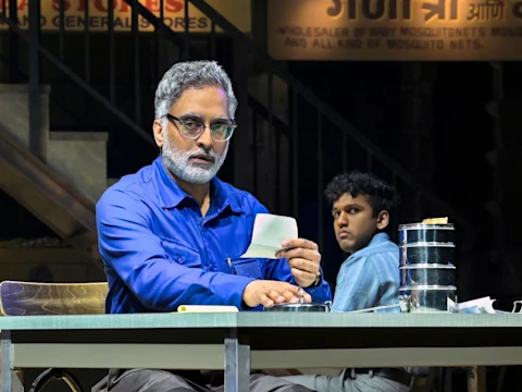 Two men sit at a desk; the man in front with gray hair and beard holds a piece of paper and looks forward, while the man behind him looks sideways. Metal containers are on the table.
