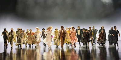 A large group of people in period costumes rushes forward on a stage under dramatic lighting.