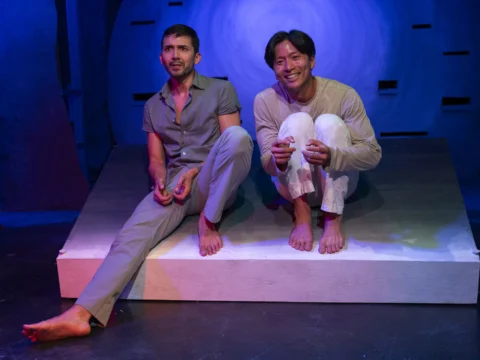 Two men sit on a slanted white platform in a dimly-lit space with blue and purple lighting. One man is dressed in grey, and the other in white. Both are barefoot and appear to be engaged in conversation.