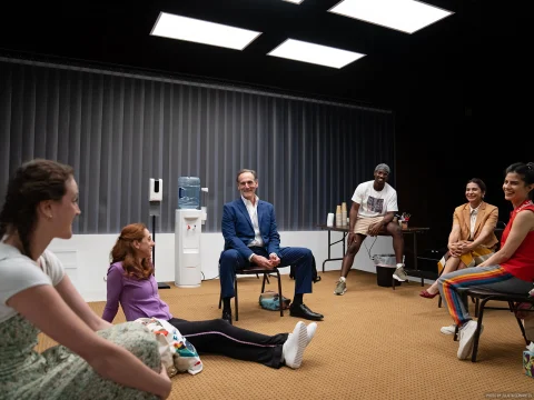 A group of people sit in a circle in a brightly lit room. Some are sitting on chairs, while others are seated on the floor. There is a water dispenser and cups in the background.