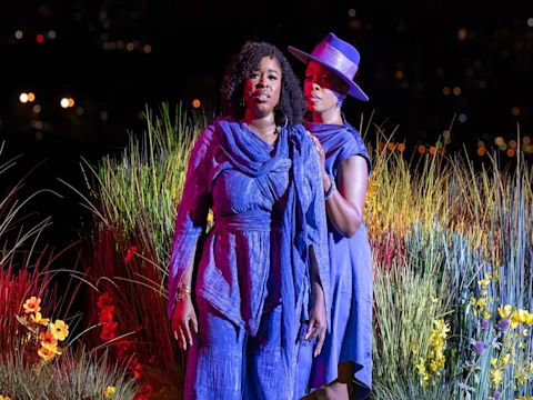 Two women in blue outfits stand among colorful flowers and tall grass at night; one stands in front while the other adjusts her scarf.