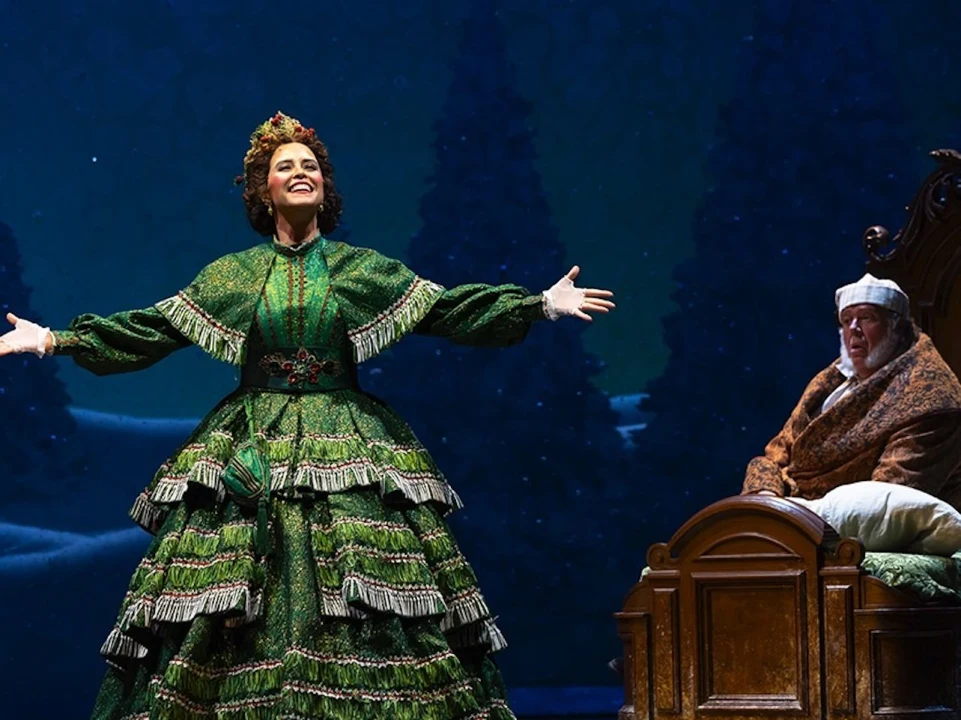 A performer in a green dress with extended arms stands beside another person sitting in a chair on stage, set against a backdrop of snow and pine trees.