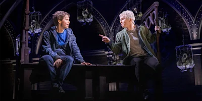 Two actors sit on a wooden structure on stage, engaged in conversation, with gothic arches and hanging lanterns in the background.