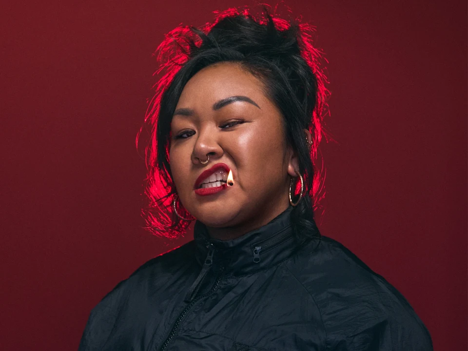 A person in a black jacket with hoop earrings and facial piercings bares their teeth, gripping a matchstick between them, against a red background.