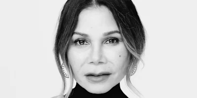 Black and white portrait of a woman with shoulder-length hair, wearing earrings and a high-neck top, looking directly at the camera against a plain background.