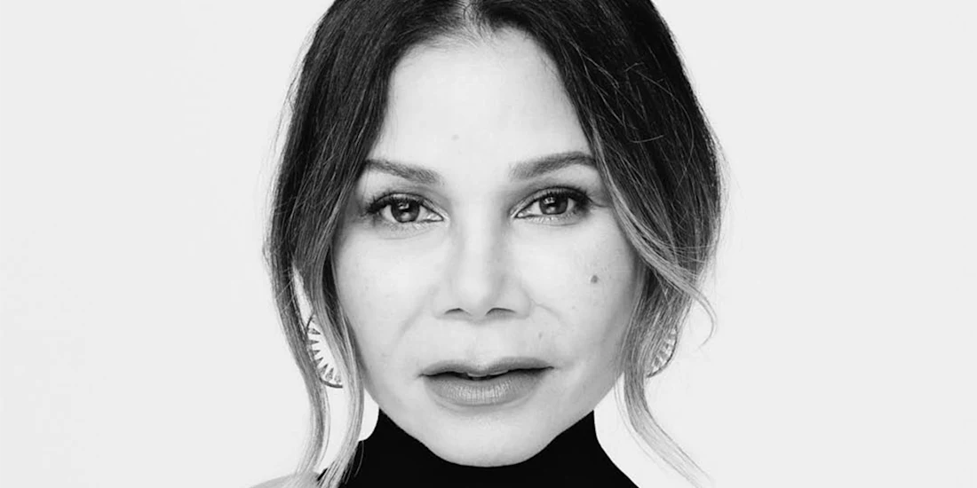 Black and white portrait of a woman with shoulder-length hair, wearing earrings and a high-neck top, looking directly at the camera against a plain background.