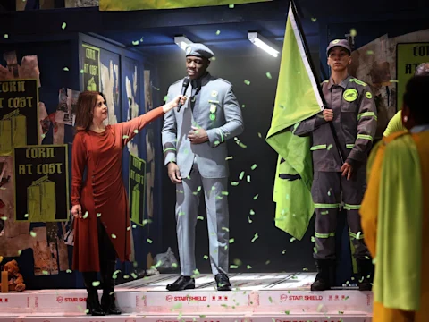 A woman in a red dress holds a microphone toward a man in a gray suit and hat, while another man in a gray uniform holds a large green flag; green confetti falls around them.