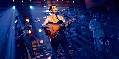 Three musicians perform on stage under blue and orange lights; the central figure sings and plays guitar, with a bassist and violinist in the background.