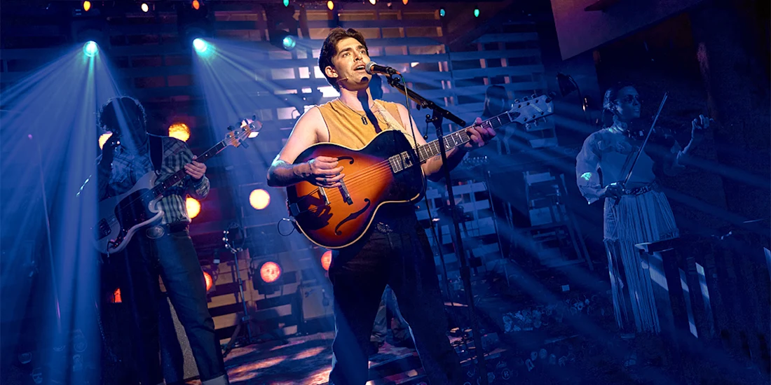 Three musicians perform on stage under blue and orange lights; the central figure sings and plays guitar, with a bassist and violinist in the background.