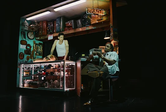 Two men are in a dimly lit pawn shop; one stands behind a glass display counter, while the other sits on a chair, gesturing as they talk.