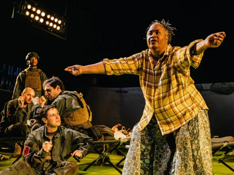 A person stands with outstretched arms, speaking animatedly, while a group of individuals in military attire sit and listen in a theatrical setting.