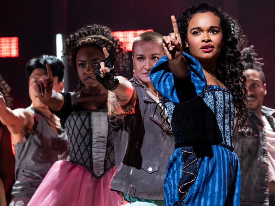 A group of performers in colorful costumes extends one arm forward with fingers pointing. The lead performer wears a blue dress, and the background is lit with red stage lights.