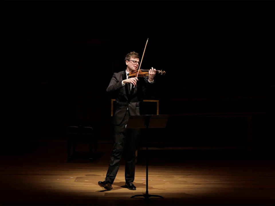 A person in a suit plays the violin on a dimly lit stage, standing in a spotlight with a music stand in front of them.