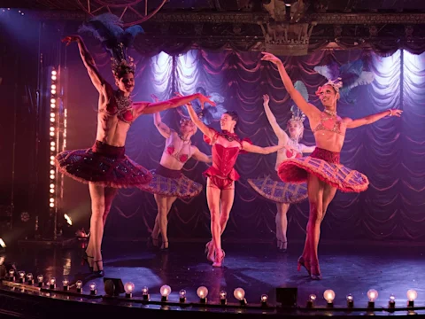 Four ballet dancers wearing colorful tutus and feathered headdresses perform on stage in front of a red, pleated curtain under stage lights.