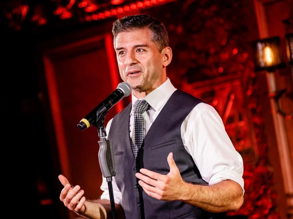A man in a white shirt, patterned tie, and dark vest speaks into a microphone on stage, with red lighting and ornate decor in the background.