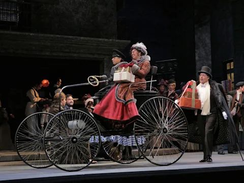 A stage scene with actors in period costumes; two people sit in a vintage car holding packages while others stand nearby, set against a city backdrop.