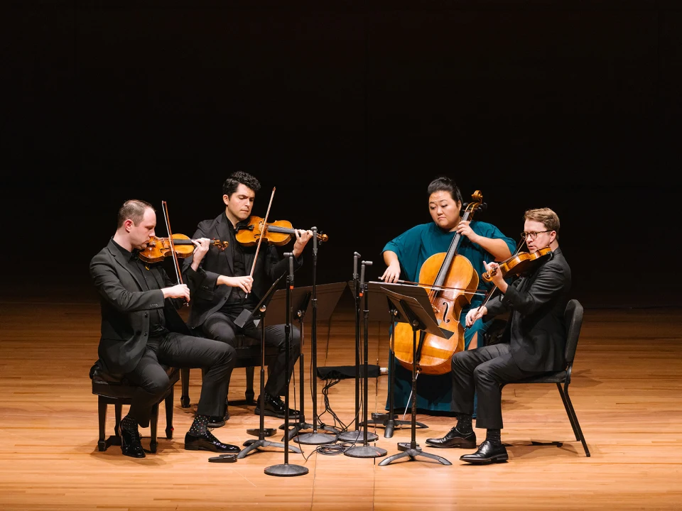 A quartet of musicians, dressed in formal attire, perform on a stage. They play string instruments: two violins, a cello, and a viola. Music stands and microphones are positioned in front of them.