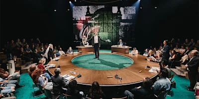 A person stands speaking at the center of a round table surrounded by seated people, with two desks and a large audience in a dimly lit room.