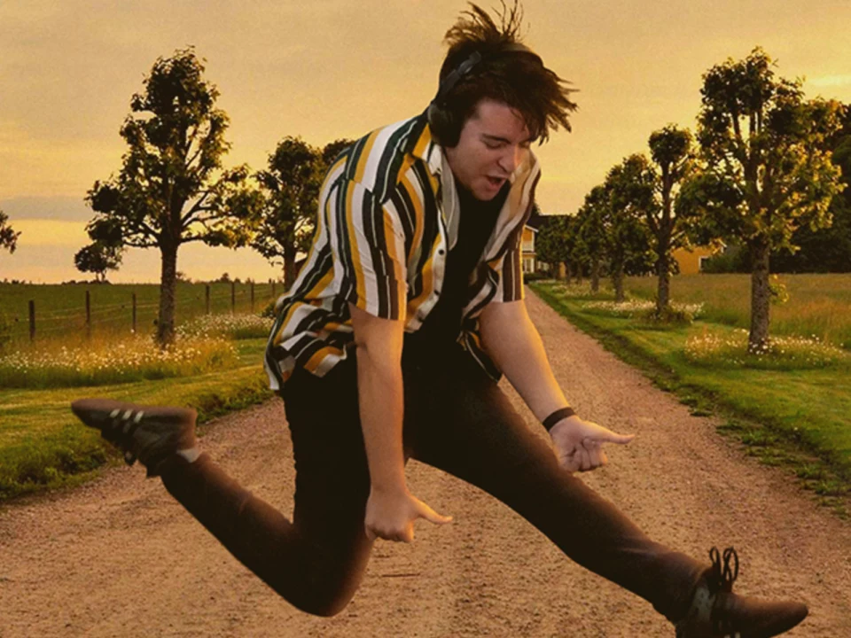 A person wearing headphones and a striped shirt jumps energetically on a rural dirt path lined with trees, with a house in the background and soft evening light.