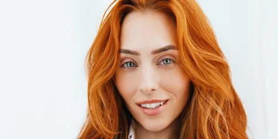 A woman with long red hair and light eyes is standing in front of a plain light background, looking at the camera and smiling subtly.