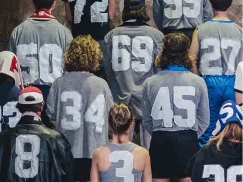 A group of people stands with their backs to the camera, wearing gray shirts displaying large white numbers.