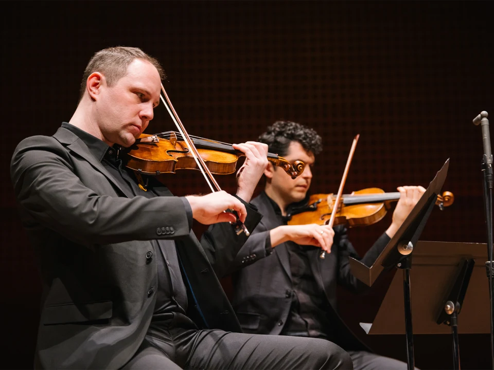 Two violinists seated, playing violins in a live performance. Both are dressed in black suits and focused on their instruments. A music stand is visible in front of them.