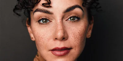 A person with curly dark hair, freckles, and well-defined eyebrows looks directly at the camera against a dark background.