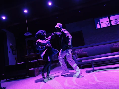 Two people dance together in a living room under purple lighting, surrounded by modern furniture and decor.
