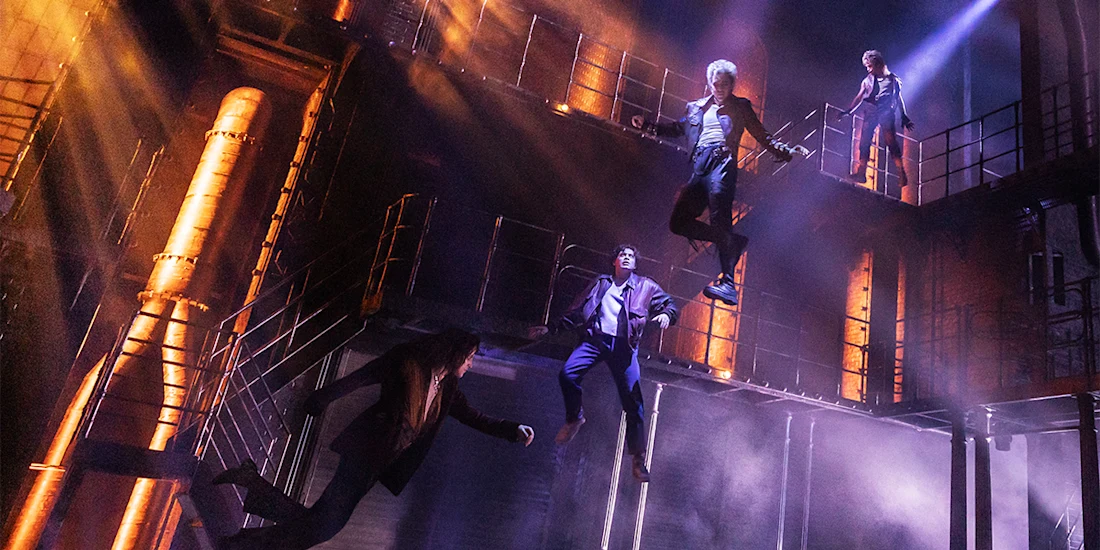 Four people in dramatic poses appear suspended in mid-air on a dimly lit industrial stage with metal walkways and orange lighting.