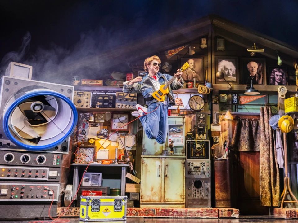 Caden Brauch as Marty McFly performed in 80s attire jumps mid-air while playing an electric guitar on a cluttered stage filled with retro electronics and large speakers.