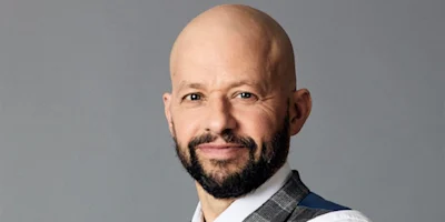 A bald man with a short beard and mustache, wearing a white shirt and vest, posing in front of a plain gray background.