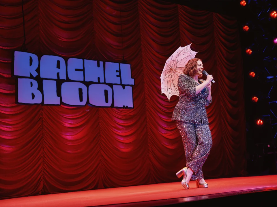 Production of photo of Rachel Bloom: Death, Let Me Do My Show in Chicago with the performer holding a parasol, standing in front of a red curtain.