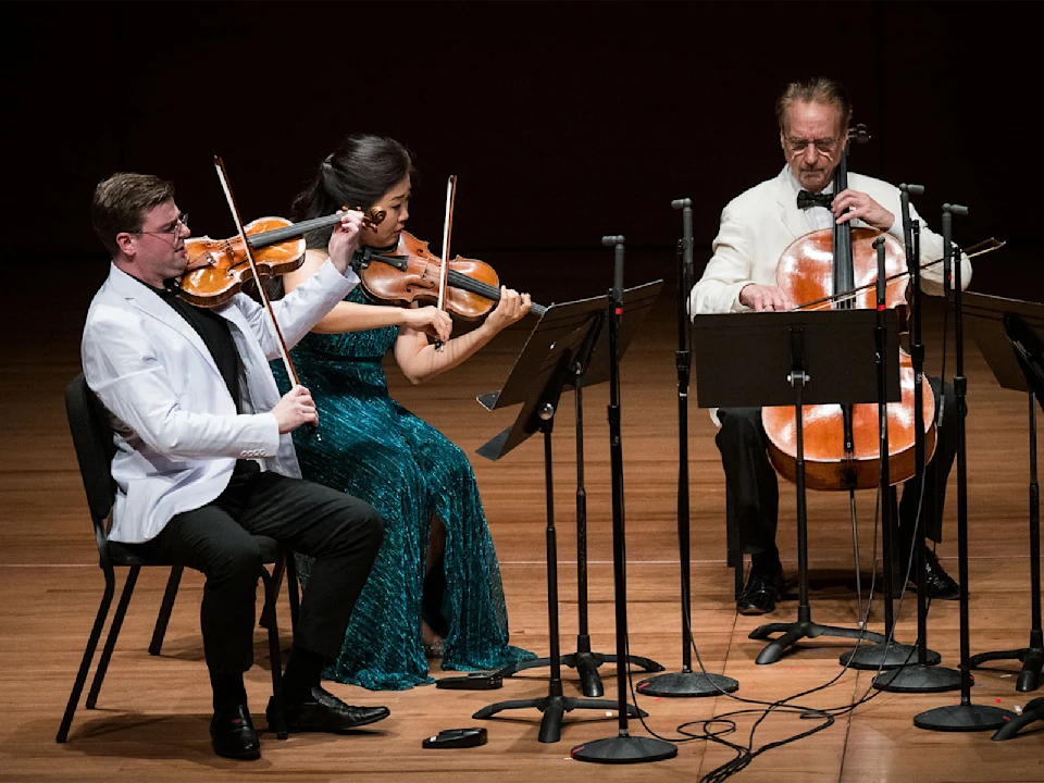 Three musicians perform on stage: two violinists and one cellist, seated with music stands and microphones in front of them.