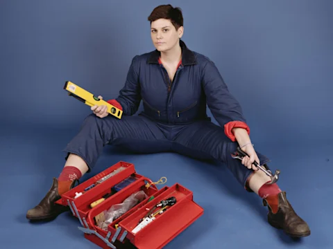 A person in a blue coverall sits on the floor with legs apart, holding a spirit level and a hammer, next to an open red toolbox filled with tools.
