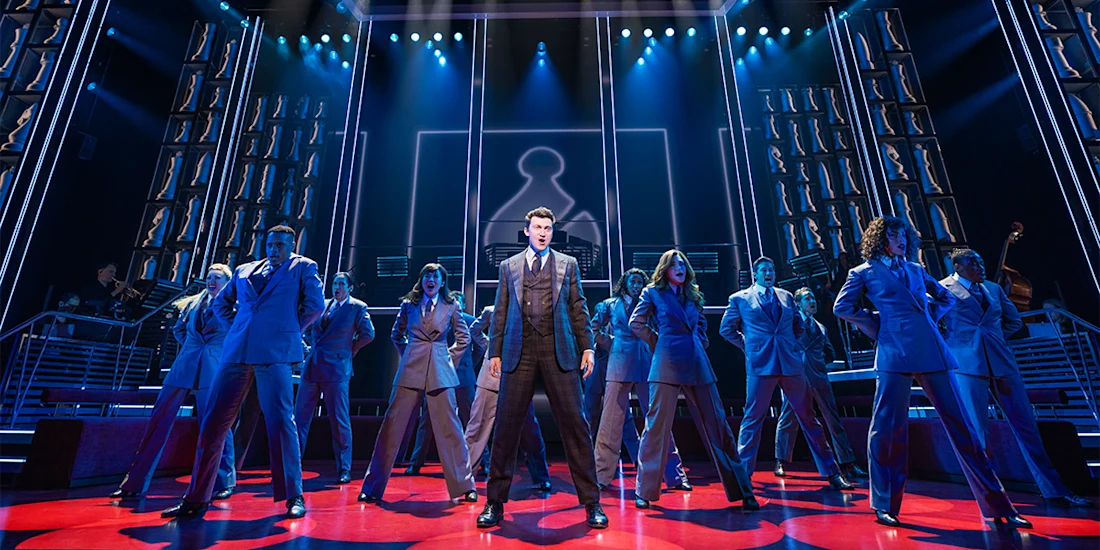 A group of performers in suits stand on stage under blue lighting, with one person in the center spotlight and a geometric backdrop behind them.