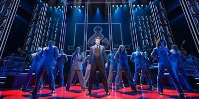 A group of performers in suits stand on stage under blue lighting, with one person in the center spotlight and a geometric backdrop behind them.