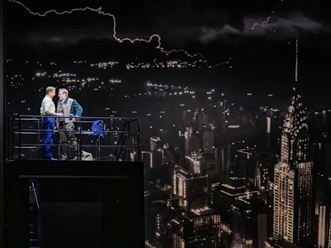 Two men stand closely together on a balcony above a cityscape at night, with illuminated skyscrapers and a dark, cloudy sky in the background.